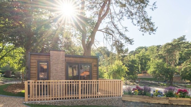 En liten, modern byggnad i stugstil med en träveranda och stenväggar, omgiven av en välskött trädgård och träd.