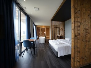 A modern hotel room with a bed, a desk with a chair, a bathtub, and large windows. - Wellness Mode 2 (Olburgen)