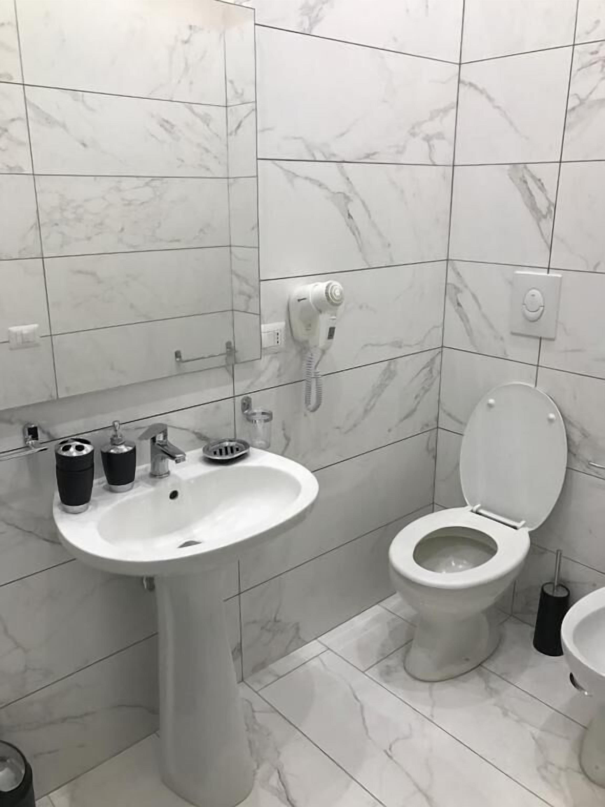 A bathroom with a white sink, toilet, and bidet, marble tiles, and a hairdryer on the wall.