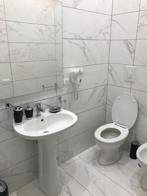 Un baño con lavamanos blanco, inodoro y bidé, baldosas de mármol y un secador de pelo en la pared.