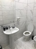 Un baño con lavamanos blanco, inodoro y bidé, baldosas de mármol y un secador de pelo en la pared.
