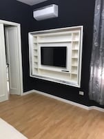 Una habitación con un televisor montado, un equipo de aire acondicionado blanco y una cortina estampada.