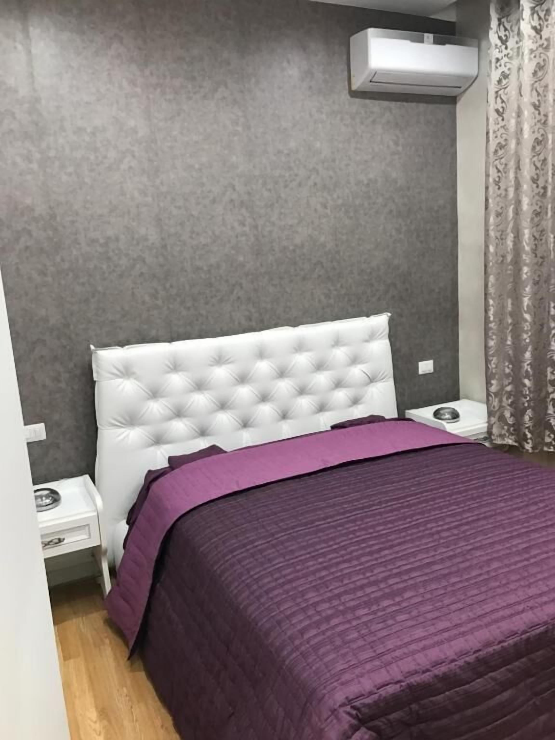 Un dormitorio con cabecera capitonada, colcha morada, mesita de noche y aire acondicionado instalado en la pared.