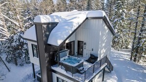 A modern cabin with a hot tub, deck, and a snowy forest backdrop.