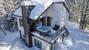 A modern cabin with a hot tub, deck, and a snowy forest backdrop.