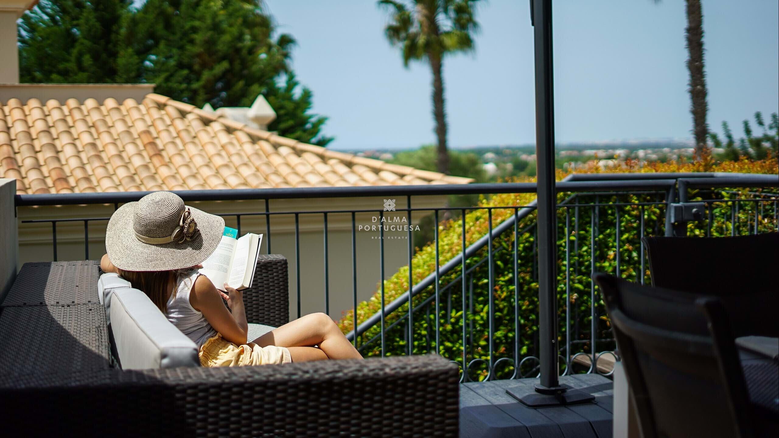 A person reading on a balcony with a view of a tiled roof and greenery.