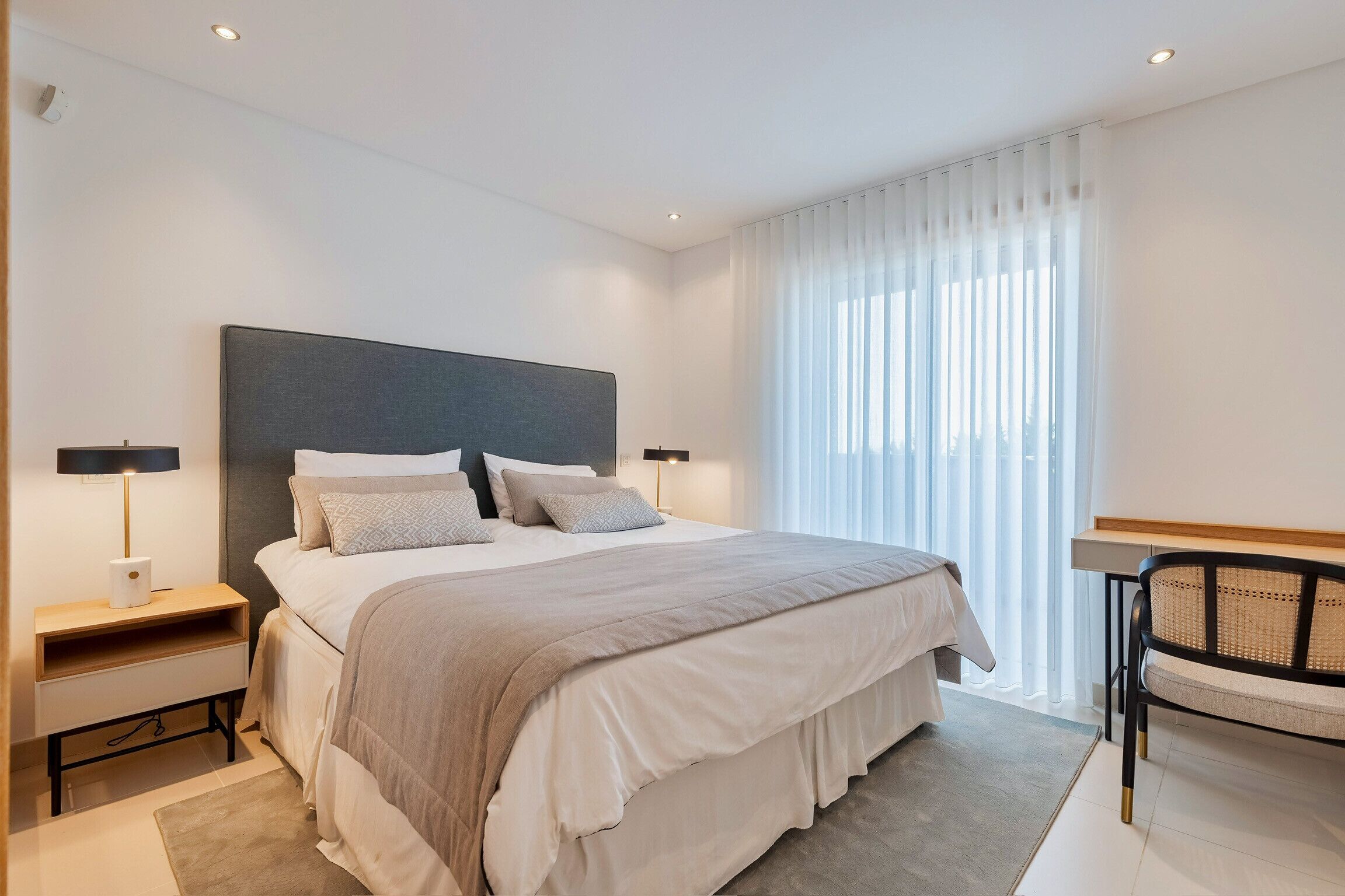 A modern bedroom with a large bed, bedside tables, a chair, and a window with sheer curtains.