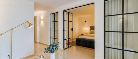 A modern hotel room with a glass partition, a round coffee table with a vase, a floor lamp, and a bed with a nightstand.