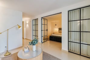 Ein modernes Hotelzimmer mit Glastrennwand, rundem Couchtisch mit Vase, Stehlampe und einem Bett mit Nachttisch.
