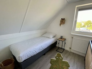 Ein kleines Zimmer mit einem Einzelbett, schrägem Dach, Heizkörper, Fenster mit Blick und einem aufgehängten Tierkopf-Dekoration.