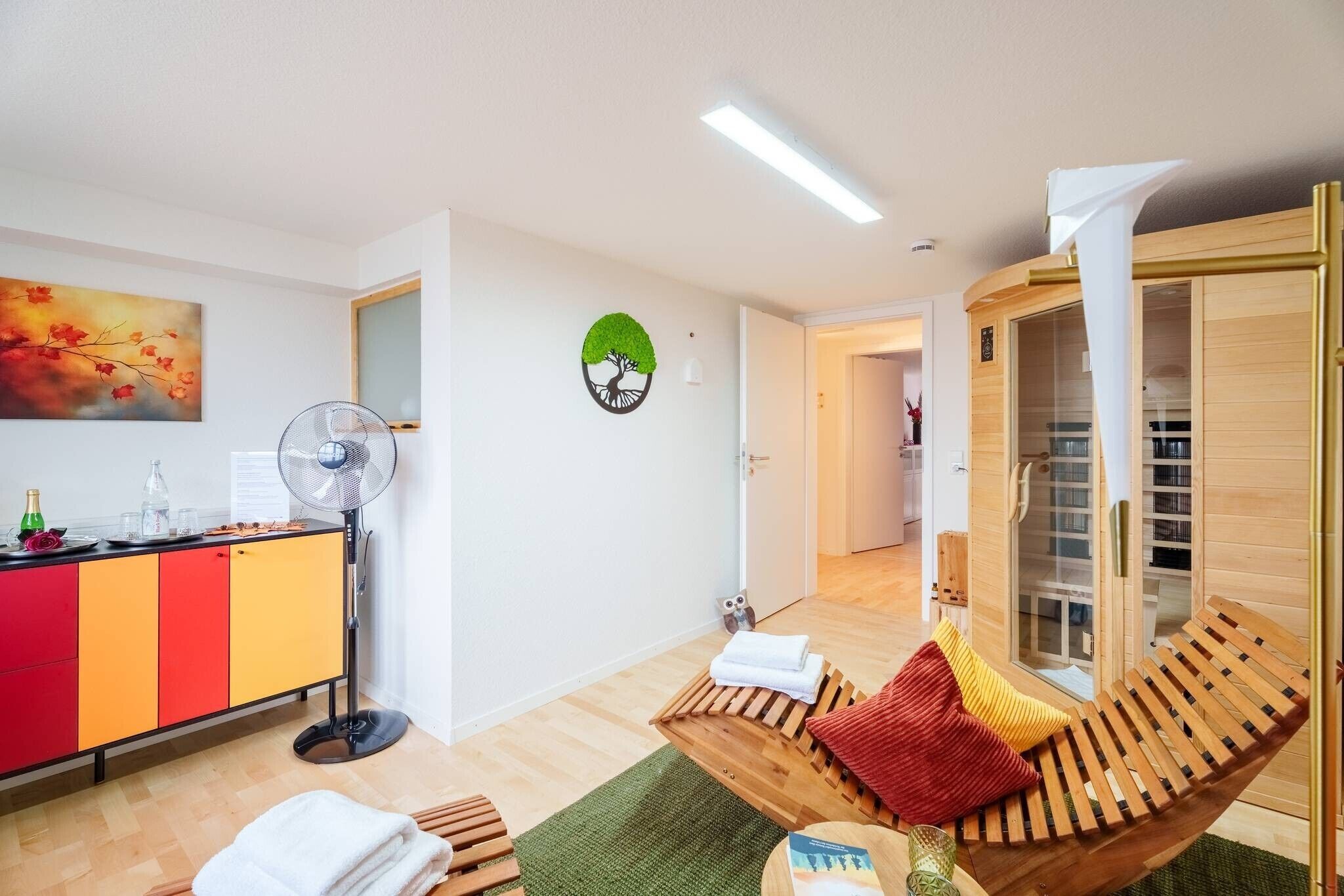 A room with a wooden bench, a fan, a cabinet with red and yellow doors, a green rug, and a wall clock.