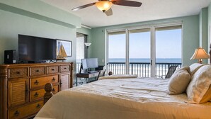 Ein Schlafzimmer mit einem Bett, einem Fernseher, einem Schminktisch, einem Schreibtisch mit Computer und einem Balkon mit Meerblick.