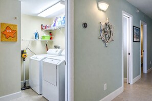 A laundry area with a washing machine, dryer, and a vacuum cleaner. There is a wall-mounted light fixture and a decorative ship's wheel. - Oceanfront Luxury Condo Million-Dollar Views (Ponce Inlet)