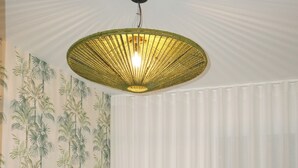 Et hotelværelse med en seng, et grønt sengegavl, en væg med tropisk tapet, et stort vindue med hvide gardiner og en hængende lampe.
