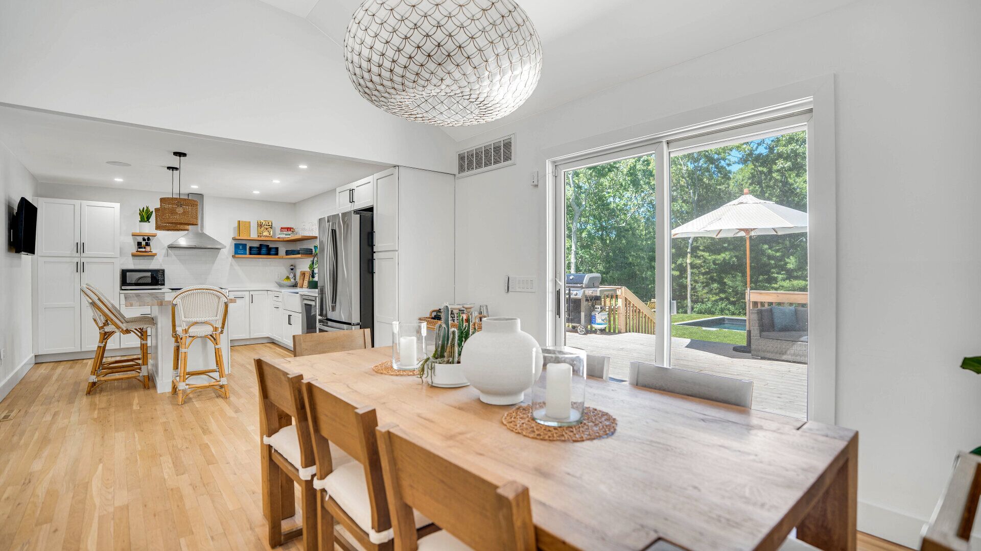 A modern kitchen with white cabinets, a wooden dining table, wicker chairs, and a large window overlooking a backyard.