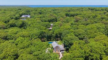 An aerial view of a property surrounded by dense forest, featuring a large house with a swimming pool and a smaller building.
