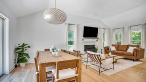A spacious living room with a wooden dining table, a brown sofa, and a large TV mounted on the wall.