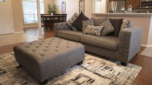 Ein Wohnzimmer mit grauem Sofa, gemusterten Kissen, einem gesteppten Ottomanen und einem Teppich mit Muster.