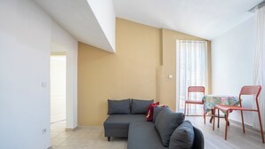 Una sala de estar con un sofá seccional gris, una mesa de comedor con sillas rojas y un ventanal con cortinas ligeras.