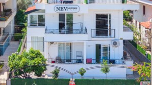 Un edificio de varios pisos con balcones, un letrero que dice 'NEV FETHYVE Rooms & Sales', y un paisaje con colinas al fondo.