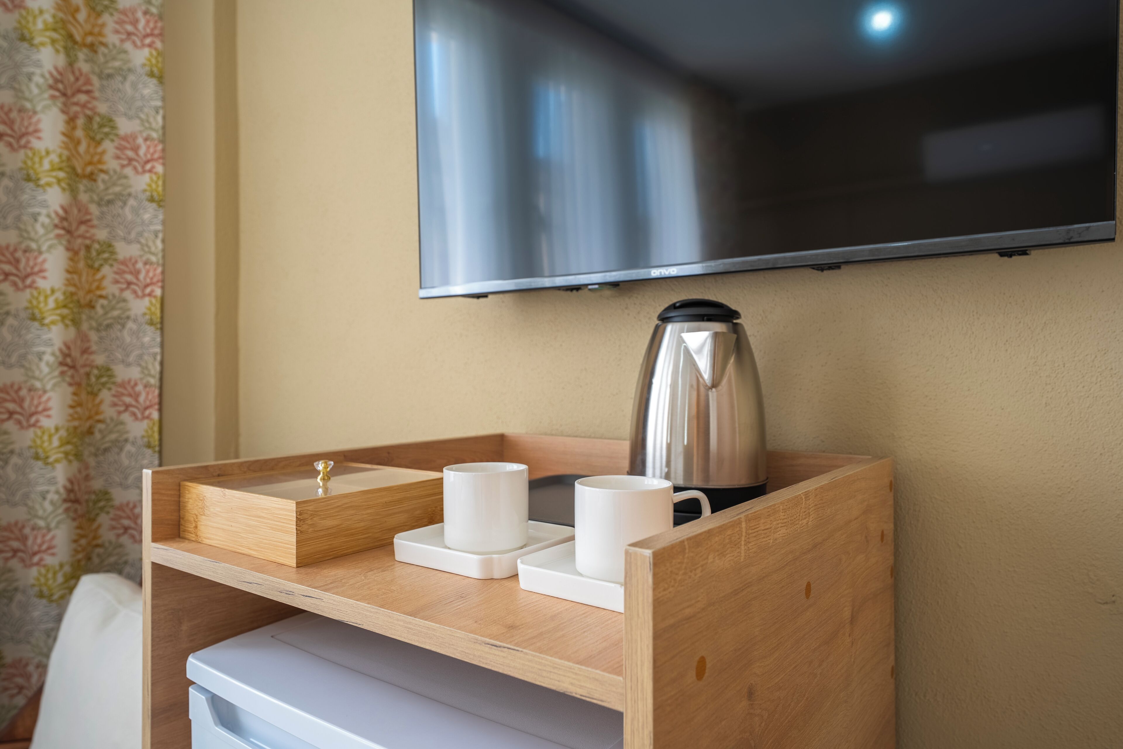 A wooden bedside table with a kettle, two white cups, and a tray. A television is mounted on the wall above.