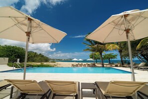 Een zwembadterras met ligbedden en parasols, een heldere blauwe lucht en uitzicht op het strand en de zee.