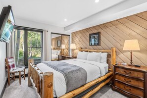 A bedroom with a wooden bed, bedside tables, a TV, and a view of greenery. - The Guilded Antler | Ski Access, Hot Tub & Pool (Beaver Creek)