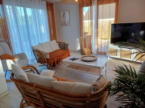 Uma sala de estar com sofá, cadeiras, mesa de centro, televisão e uma janela grande com cortinas.