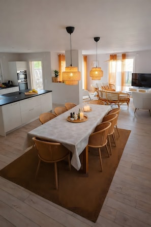 Una cocina moderna con armarios blancos, una zona de comedor con una mesa larga y sillas de mimbre, y una sala de estar con sofá y televisor.