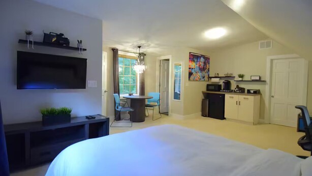 A bedroom with a large bed, a TV mounted on the wall, a desk with a chair, a dining area with a chandelier, and a kitchen area. - Backwoods Studio: 11.6ac Forest (Noblesville)
