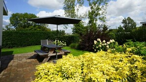 A patio with a table and umbrella, surrounded by yellow flowering plants and a hedge. - Cube Maximaal 4 (Halfweg)