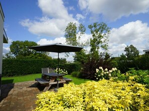 Eine Terrasse mit Tisch und Sonnenschirm, umgeben von gelb blühenden Pflanzen und einem Heckenstreifen.