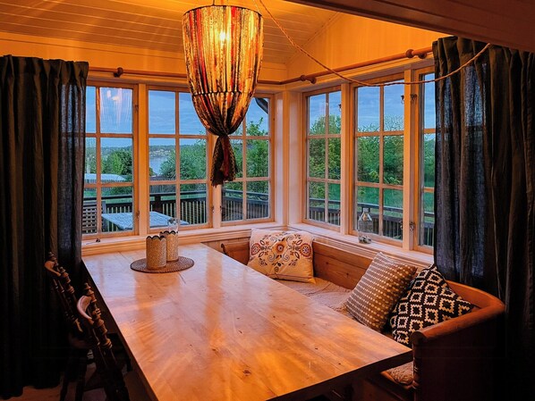 Ein Zimmer mit Holztisch, einer Bank und großen Fenstern mit Blick auf viel Grün.