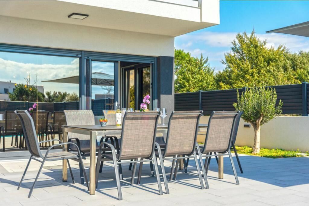 Un espacio al aire libre para comer con una mesa, sillas, un árbol pequeño y un edificio con ventanas grandes.