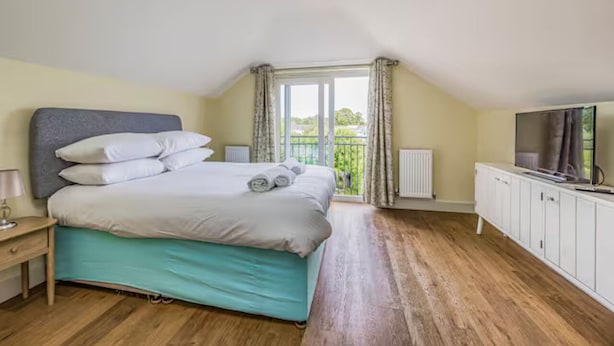 Ein Schlafzimmer mit Bett, Nachttisch, Fernseher, Heizkörper und einem Balkon mit Vorhängen.