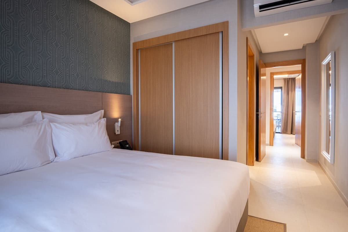 A hotel room with a large bed, a wardrobe, a bedside table with a lamp, and a view of the hallway.