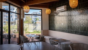 An indoor dining area with tables and chairs, a large window with a view of buildings, and hanging woven light fixtures.