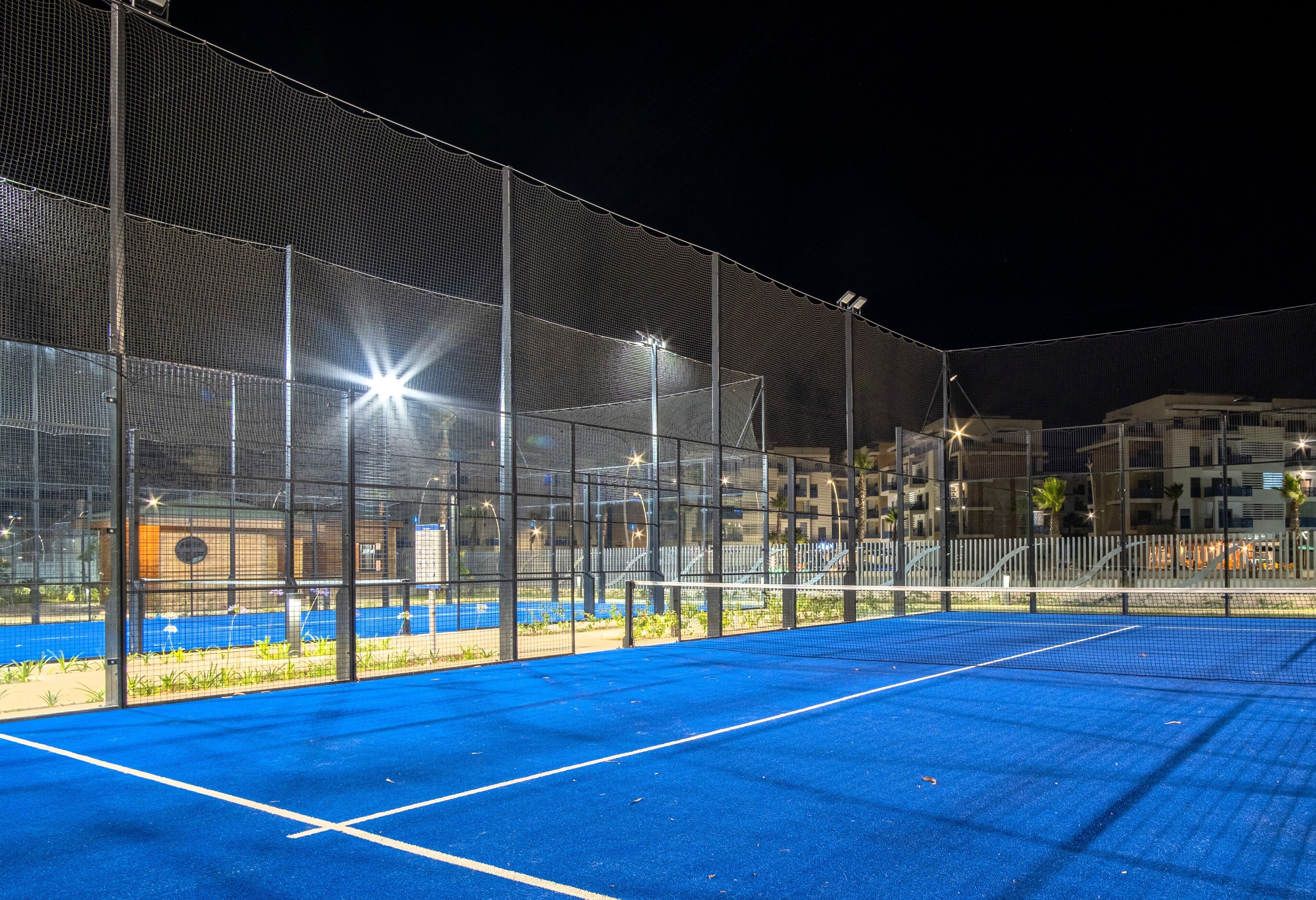 An outdoor tennis court with a net, surrounded by a fence, and illuminated by overhead lights.