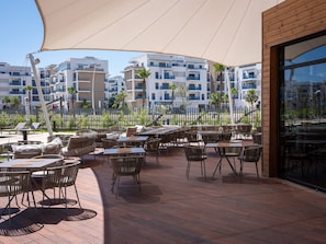 An outdoor patio with tables and chairs, a wooden deck, and a view of modern buildings in the background.