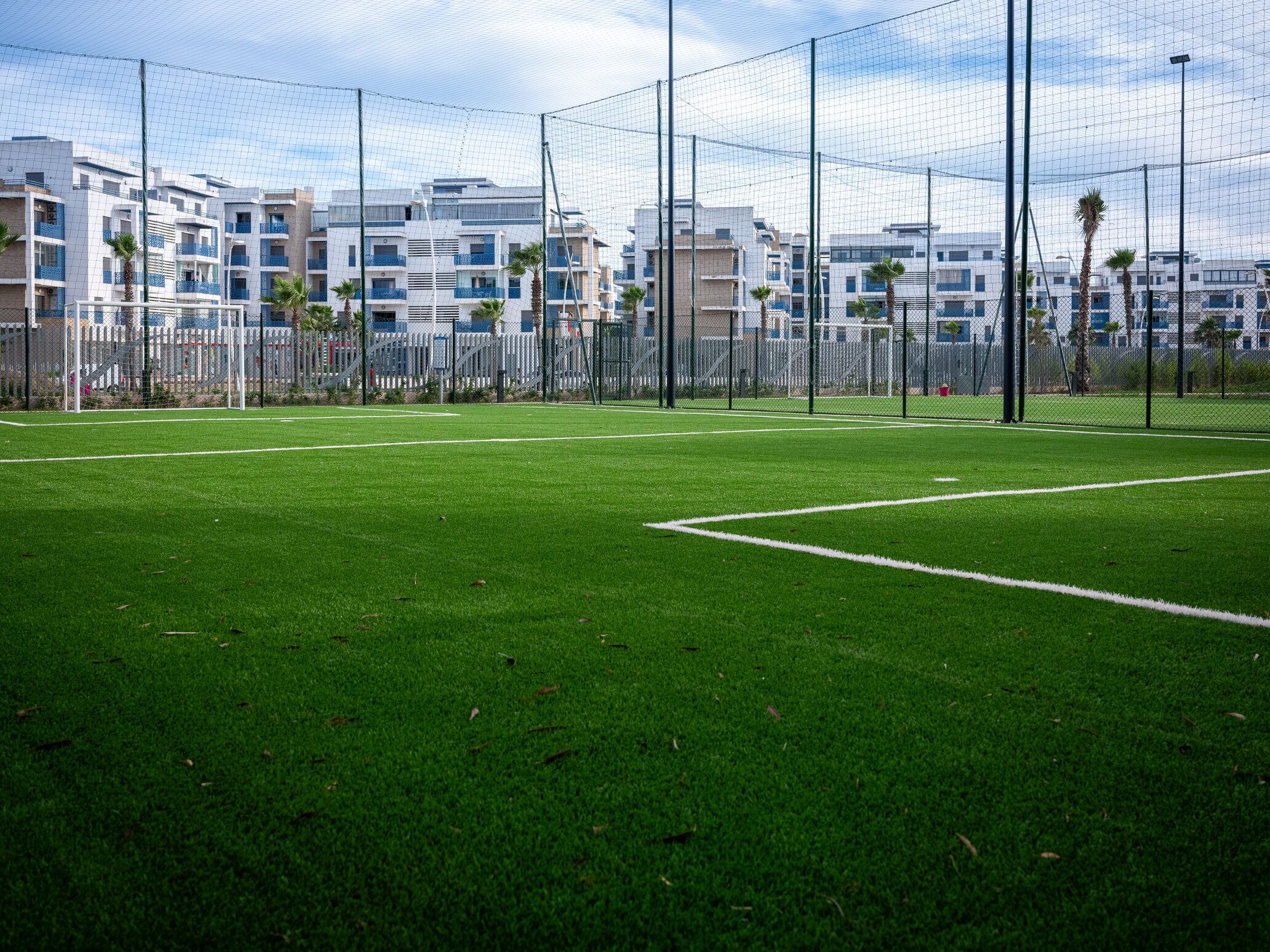 Un terrain de sport avec des poteaux de but, entouré d’une clôture et bordé de bâtiments en arrière-plan.