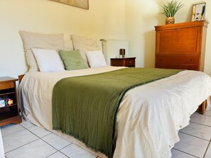 Ein ordentlich bezogenes Bett mit grüner Tagesdecke, ein Nachttisch aus Holz mit Lampe und ein Holzschrank mit Pflanze darauf.