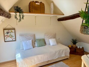 Een knus slaapkamer met een bed, kussens, een hangplant, een houten kist en een ingelijste foto aan de muur.