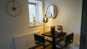 Un coin repas comprenant une table, deux chaises, un miroir rond, une horloge murale et une fenêtre avec des stores.