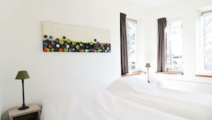 A hotel room with a bed, two bedside lamps, a window with curtains, and a wall art piece. - Groepsaccommodatie 18 Otterlo (Otterlo)