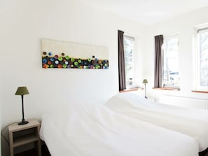 A hotel room with a bed, two bedside lamps, a window with curtains, and a wall art piece. - Groepsaccommodatie 18 Otterlo (Otterlo)