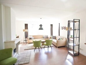 A living room with a sofa, two chairs, a floor lamp, a bookshelf, and a fireplace. - Groepsaccommodatie 18 Otterlo (Otterlo)