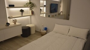 A bedroom with a bed, bedside tables, a nightstand, a small stool, a wall-mounted TV, and shelves with decorative items. - Flisvos Beach Retreat (ΑΘΗΝΑ)