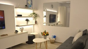 A modern living room with a grey sofa, a white table, a TV, and a mirror. - Flisvos Beach Retreat (ΑΘΗΝΑ)
