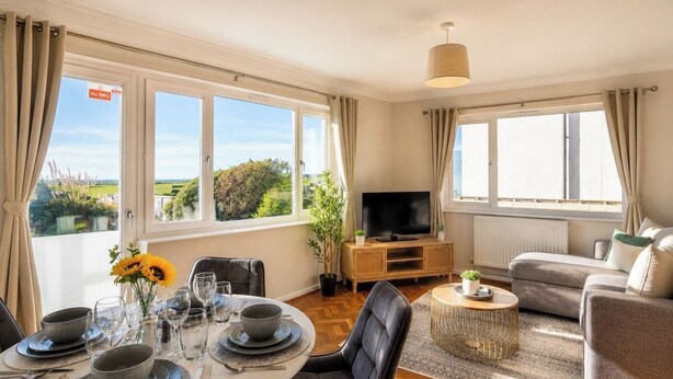 A well-lit room with a dining table set for four, a sofa, a TV, and a view of greenery outside. - Amazing Location - Free Parking - Beach Opposite! (Bournemouth)