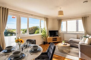 Une pièce bien éclairée, avec une table à manger dressée pour quatre personnes, un canapé, un téléviseur et une vue sur la verdure à l’extérieur.